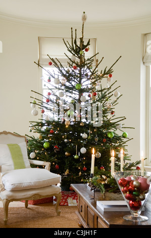 Geschmückter Weihnachtsbaum mit Kugeln im Wohnzimmer mit Sessel und Kerzen Stockfoto