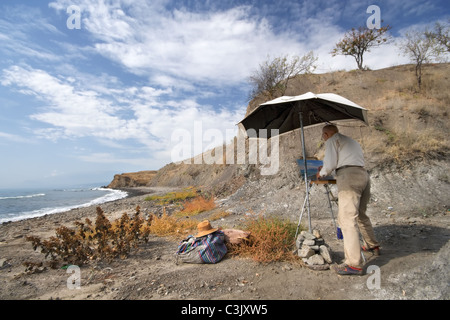 Maler malt Bild an der Meeresküste im Herbst. In der Nähe gibt es Büsche, seinen Hut und Tasche. Im Hintergrund Meer und Himmel. Stockfoto