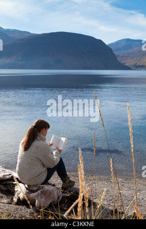 Frau sitzt am Fjord und Buch Stockfoto