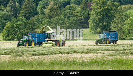 2 John Deere-Traktoren und West-Anhänger arbeiten und fahren auf landwirtschaftlichen Feldern mit Claas-Feldhäcksler, beladen von geschnittenem Gras (Silage) - Yorkshire, England. Stockfoto