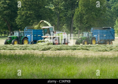 2 John Deere-Traktoren und West-Anhänger arbeiten und fahren auf landwirtschaftlichen Feldern mit Claas-Feldhäcksler, beladen von geschnittenem Gras (Silage) - Yorkshire, England. Stockfoto