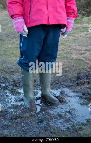Kleines Kind (Mädchen) in grünen wellington Stiefeln, rosa wasserdichter Mantel, Handschuhe & blaue Hosen stehen in Schlamm oder schlammigen Pfütze (Beine & Körper) - England. Stockfoto