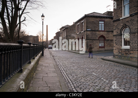 Albert Terrace im historischen Dorf Saltaire (Ende terrassenförmig angelegte Grundstücke, viktorianische Häuser, Steinbauten auf der Straße, Mühlkamin) - Bradford, Yorkshire, UK. Stockfoto