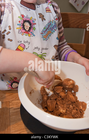 Junge Mädchen Kuchen backen, rühren Zutaten (Butter & brauner Zucker) in Schüssel mit Holzlöffel & Tracy Becher Schürze tragen - Yorkshire, England Stockfoto