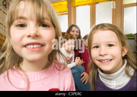 Deutschland, zwei Mädchen (3-5) im Vordergrund, die auf der Suche in Kamera, weibliche Kindergärtnerin mit Kindern im Hintergrund Stockfoto