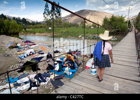 Peru, Otuzco, Indianerin mit typischen Hut Überquerung Hängebrücke. Hintergrund: Menschen, die Wäsche im Fluss waschen. Stockfoto