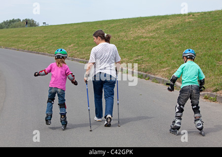 eine Frau mittleren Alters, die ihre zwei kleinen Kinder Skaten neben ihr tun Nordic Walking Stockfoto