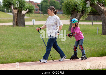 eine Frau mittleren Alters, Nordic Walking, ziehen ihre junge Tochter auf Schlittschuhen hinter ihr zu tun Stockfoto