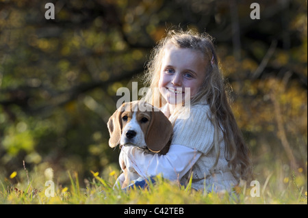 Beagle (Canis Lupus Familiaris). Kleines Mädchen mit einem Welpen auf dem Arm sitzen auf einer Wiese. Deutschland. Stockfoto