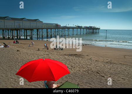 Die Grand Pier und Urlauber sitzen unter einem roten Sonnenschirm in Teignmouth, Devon, England, UK Stockfoto