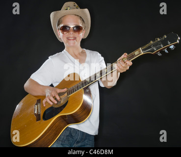 Der junge Schuljunge spielt Akustikgitarre, das Studio beleuchtet sein Gesicht mit Sonnenbrille und Cowboyhut Stockfoto