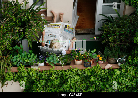 Eine ältere deutsche Frau sitzt entspannt auf ihrem Balkon Zeitunglesen umgeben von Blumentöpfe in Leipzig, Deutschland Stockfoto