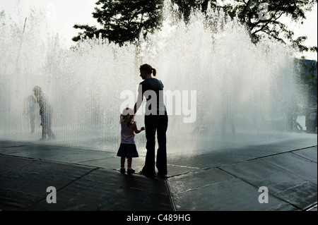 Frau und Mädchen gegen Wasser Skulptur erscheinen Zimmer Silhouetted, spielende Kinder im Brunnen außerhalb der Royal Festival Hall, auf dem Londoner Southbank. Stockfoto