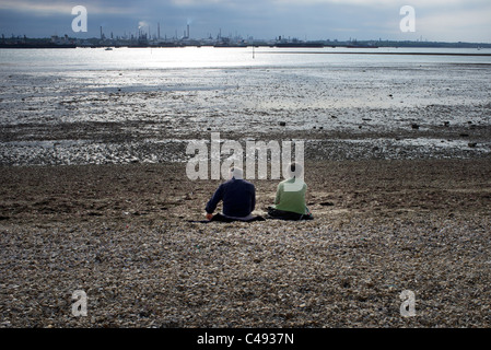 Paar sitzt auf einem Kiesstrand bei Ebbe an Southampton Wasser UK gegenüber Hythe petrochemischen Raffinerie Stockfoto