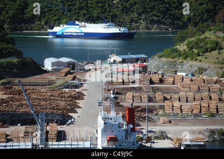 Holz aufgestapelt am Kai im Hafen Marlborough, in der Nähe von Picton, Neuseeland mit einer Bluebridge Fähre ankommen im Hintergrund. Stockfoto