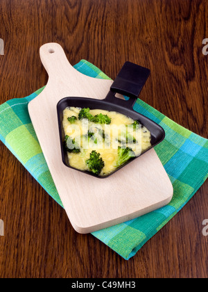 Raclette-Pfanne mit Käse und Brokkoli auf Schneidebrett Stockfoto