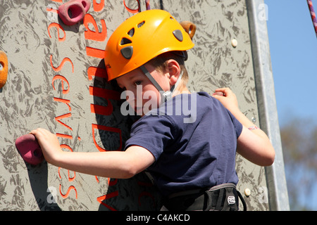 Ein kleiner Junge, lernen, wie man Aufstieg rock an einer Kletterwand außerhalb in der Sommersonne. Er ist zu seinem Gurt festgeschnallt. Stockfoto