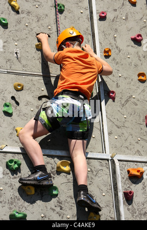 Ein kleiner Junge, lernen, wie man Aufstieg rock an einer Kletterwand außerhalb in der Sommersonne. Er ist zu seinem Gurt festgeschnallt. Stockfoto