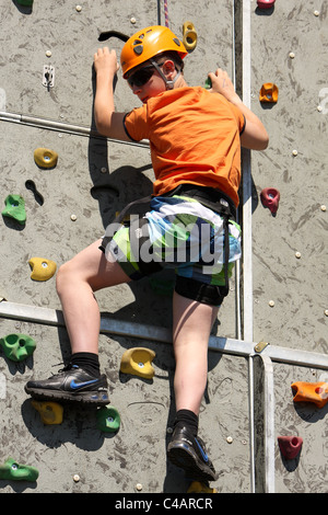 Ein kleiner Junge, lernen, wie man Aufstieg rock an einer Kletterwand außerhalb in der Sommersonne. Er ist zu seinem Gurt festgeschnallt. Stockfoto