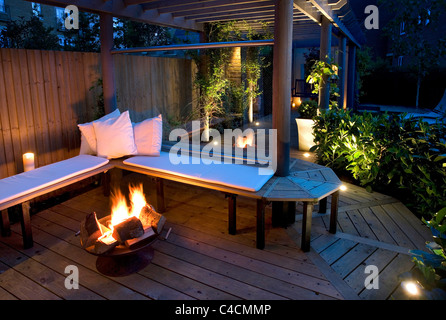 zeitgenössische moderne englische Stadtgarten mit Holzbank decking und Pergola, Wind Pause Glas und Feuerkorb in der Nacht Stockfoto