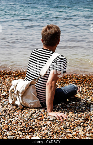 Mann mit gestreiften t-Shirt im Urlaub sitzt auf einem Kiesstrand an der Küste am Meer - Modell veröffentlicht Stockfoto