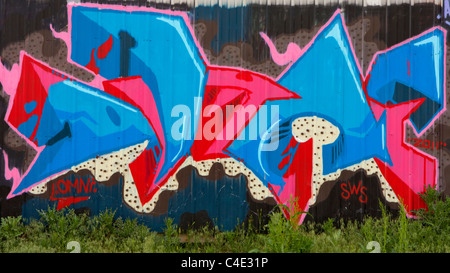 Abstrakte rosa und blau Graffiti über eine große braune gewellte Stahlwand mit Unkraut im Vordergrund. Stockfoto