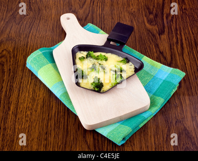 Raclette-Pfanne mit Käse und Brokkoli auf Schneidebrett Stockfoto