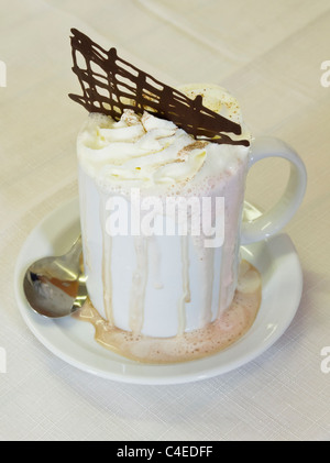 Becher der schaumigen Heißgetränk Schokolade garniert mit Schlagsahne liefen Seite in die Untertasse auf einer weißen Tischdecke Stockfoto