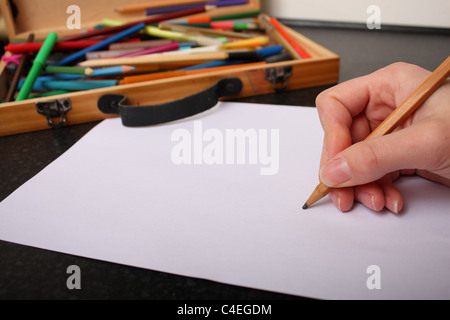Eine Hand mit einem Bleistift bereit bereit zum Zeichnen, skizzieren und Farbe auf ein Stück weißes Papier mit Farben im Hintergrund Stockfoto