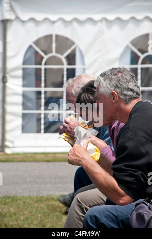 Drei Personen nebeneinander auf einer Bank sitzend Essen Cornish Pasties aus Papiertüten. Dahinter befindet sich ein weißer Zelt Pavillon Stockfoto