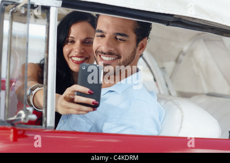 glücklicher junger Mann und Frau lächelnd während der Einnahme Snapshot mit Handy-Kamera aus roten Cabrio Oldtimer Stockfoto