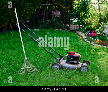 Ein gut genutzt und schmutziger, Rasenmäher mit einem Garten Rechen es gelehnt sitzt in dem verwilderten Rasen von einem Gartenhof. Stockfoto