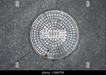 Eine alte Kanalisation Kanaldeckel umgeben durch eine Asphalt-Straße Stockfoto