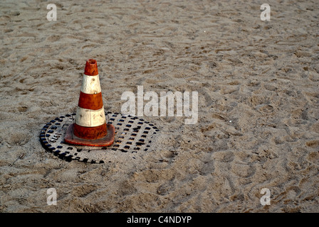 Ein orange-weiße Pylon sitzt auf einem Kanaldeckel an einem Sandstrand. Stockfoto