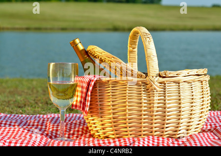 Eine Flasche Weißwein in Picknick-Korb mit Glas Wein daneben. Rot karierte Decke und Serviette mit See im Hintergrund. Stockfoto