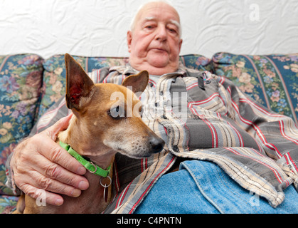 Ein pensionierter älterer Senior Mann in seine Heimat auf seiner Couch sitzen mit seinem Hund an seiner Seite. Stockfoto