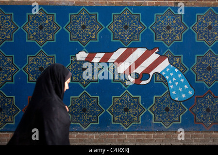 Iranische Frau, die in Teheran (oder Teheran) in der Nähe eines Graffiti mit Waffe auf der Außenwand der ehemaligen US-Botschaft „Höhle der Spionage“ im Iran spaziert Stockfoto