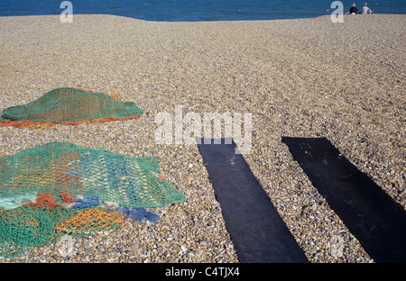 Breiten Kiesstrand mit Streifen aus schwarzem Kautschuk Matten und bunten Fischernetze und zwei entfernten Figuren sitzen in der Nähe von Meer Stockfoto