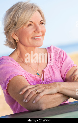 Eine attraktive elegante und glückliche senior Frau draußen sitzen aus einer Tabelle und Lächeln, hinter ihr ist ein Strand und das Meer Stockfoto