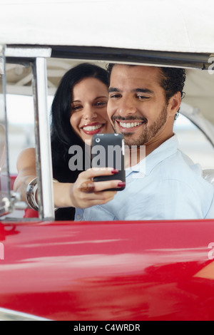glücklicher junger Mann und Frau lächelnd während der Einnahme Snapshot mit Handy-Kamera aus roten Cabrio Oldtimer Stockfoto