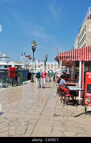 Wanderer haben einen entspannenden Spaziergang entlang der Uferstraße mit Booten und Yachten auf der einen Seite und Cafés und Restaurants auf der anderen Seite Stockfoto