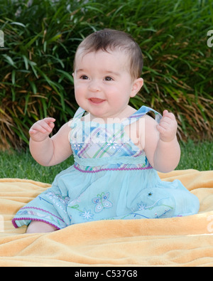 Eine acht Monate alte kaukasische lachende Baby Mädchen sitzt auf einer Decke im Freien. Stockfoto