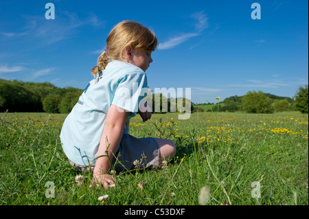 Junges Mädchen in t Shirt sitzt im Freiland auf Sommertag gelbe Butterblumen vor einem blauen Himmel betrachten. Stockfoto