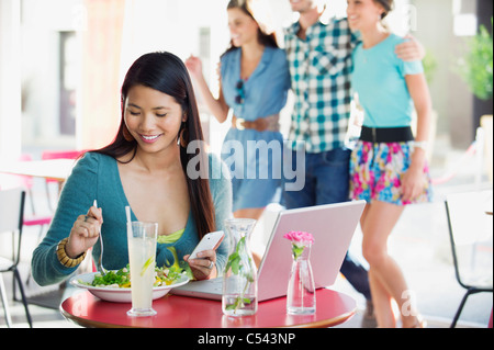 Lächelnde Frau Essen während der Verwendung mobiler mit Menschen im Hintergrund in einem restaurant Stockfoto