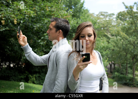 Junges Paar, das in einem Park mit Rücken gegeneinander mit dem Mobiltelefon telefoniert (von Models gestellt) Stockfoto