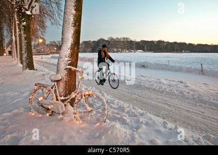 Der Niederlanden,'s-Graveland, Fahrrad dekoriert mit Weihnachtsbeleuchtung im Schnee. Frau, Radfahren. Stockfoto