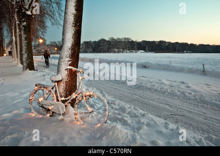 Der Niederlanden,'s-Graveland, Fahrrad dekoriert mit Weihnachtsbeleuchtung im Schnee. Frau, Radfahren. Stockfoto