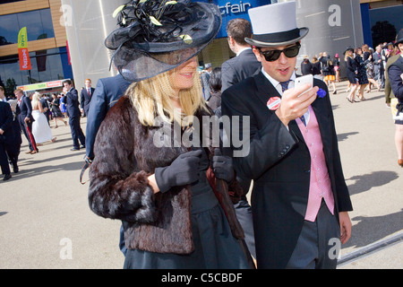 Eine Frau in einem braunen Pelzmantel und schwarzen Handschuhen in Royal Ascot geht mit einem Mann in Hut & Schwänzen & rosa Weste. Stockfoto
