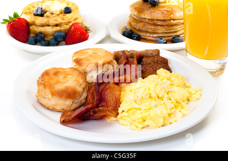 Frühstücksteller mit Rührei, Speck und Kekse Buttermilch.  Waffeln, Pfannkuchen und Orangensaft im Hintergrund. Stockfoto