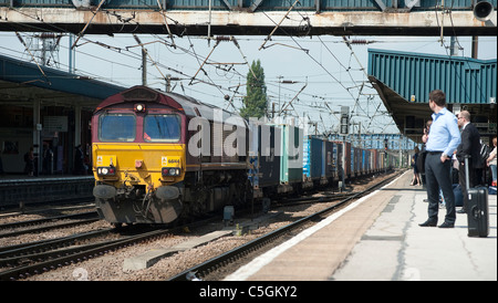 Passagiere warten auf eine Plattform als ein ein Güterzug durchläuft einen Bahnhof in England. Stockfoto